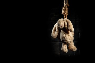 Rabbit toy, hanged on a thick braided rope on a dark background. Suicide conception.