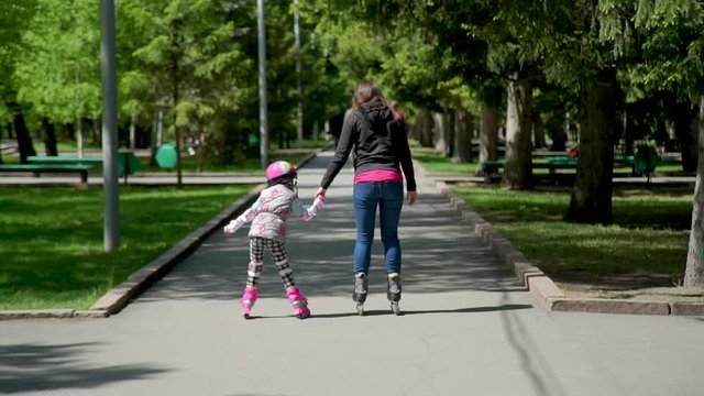 Mother with Daughter Roller Skating in a Park Together in Slow Motion. Rear View. Family Sport Activities Outdoors. People, Children, Childhood Concept