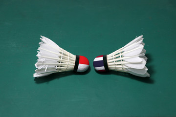 Two used shuttlecocks on green floor of Badminton court with both head each other. One head painted with Indonesia flag And one head painted with the Thai flag.