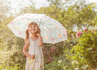 Happy little girl in garden under the summer rain with an umbrella.