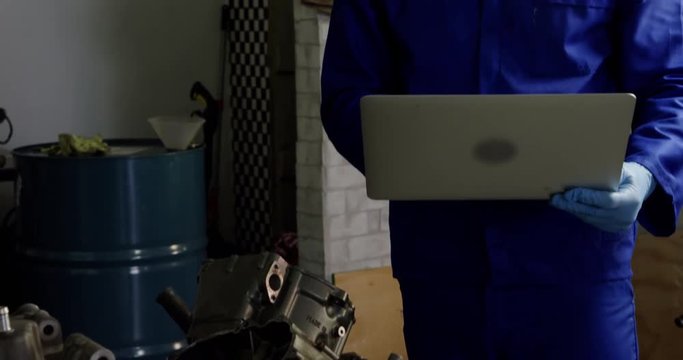 Male mechanic using laptop while repairing motorbike engine in garage.