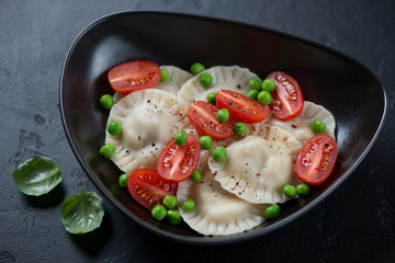 Black bowl with ravioli, cherry tomatoes and green peas, studio shot on a black stone background