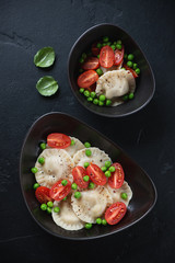 Vareniki dumplings with green peas and cherry tomatoes served in two bowls, flatlay on a black stone background