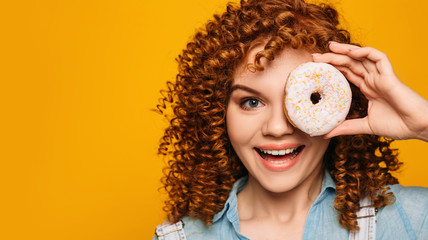 curly red-haired woman holding doughnut near face with surprise emotional face on yellow background