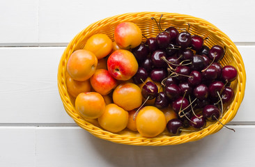  sweet cherries and ripe apricots in a yellow basket on a white wooden background
