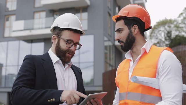 Construction manager and architect man are discussing the construction plan of the modern business center. They checks the work with a tablet. Outside.