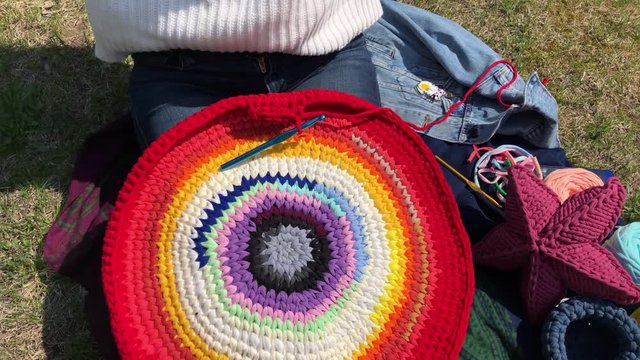 Young beautiful woman knits a rug of yarn sitting on the lawn in the park on a sunny spring day. The girl is engaged in a calm hobby on the street