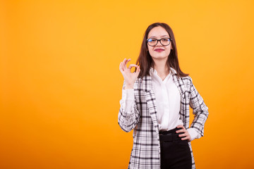 Cheerful adolescent girl with eyeglasses showing ok sign to the camera in studio over yellow background