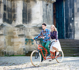Fototapeta premium Young active happy traveler couple, handsome bearded man and pretty blond woman in glasses riding together tandem bicycle along paved sidewalk on bright sunny summer morning by ancient buildings.