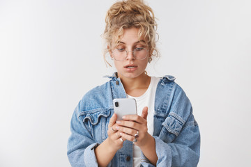 Portrait worried intense good-looking stylish modern 20s young woman blond curly messy bun wear glasses denim jacket look disturbed nervous smartphone screen reading message, checking grocery list