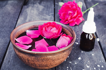 Essential oil near the bowl with red rose petals on a dark wooden background.
