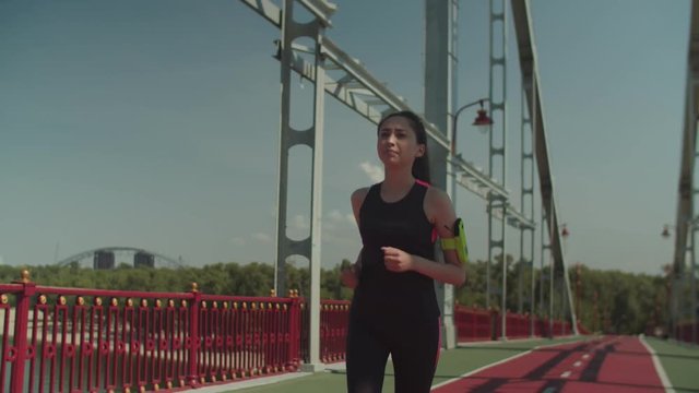 Active fitness asian woman in sporty outfit jogging along pedestrian bridge during morning workout. Chinese athlete female with armband exercising outdoors running across bridge in the city.