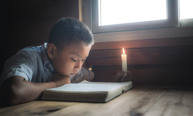 Christian boy readidng bible with light of candle on wooden table near the window at home. focus at face.