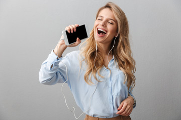 Photo of joyful blond businesswoman with long curly hair singing while listening to music with smartphone and earphones