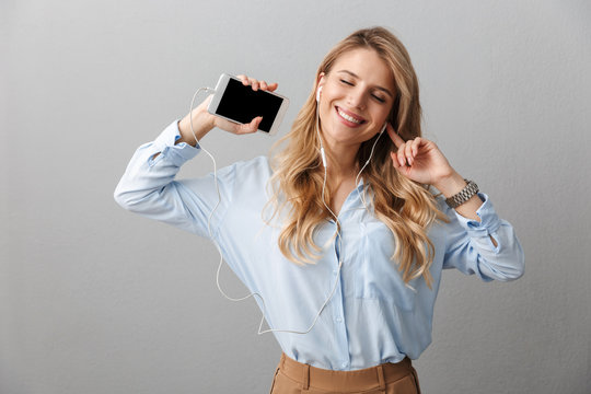 Photo of charming blond businesswoman with long curly hair rejoicing while listening to music with smartphone and earphones