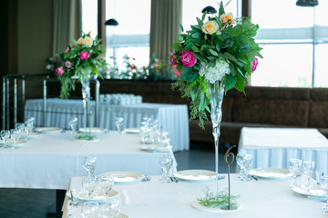 Beautiful table setting with crockery and flower arrangement in a vase on a high stem for a party, wedding reception or other festive event. Glassware and cutlery for catered event dinner