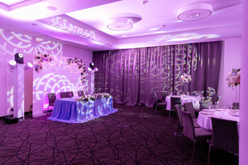 Wedding banquet in blue and white tones. Dinner table for newlyweds decorated with greenery and long cloth. A long flower arrangement on the table and a floral decor on a white wooden wall