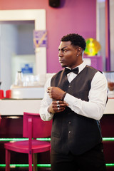 Young handsome african man wearing white shirt, black vest and bow tie posed against bar counter at night club.