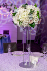Beautiful table setting with crockery and white flower arrangement in a vase on a high stem for a party, wedding reception or other festive event. Glassware and cutlery for catered event dinner