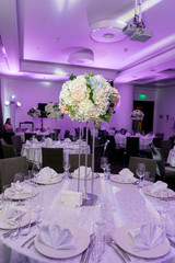 Beautiful table setting with crockery and white flower arrangement in a vase on a high stem for a party, wedding reception or other festive event. Glassware and cutlery for catered event dinner
