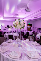 Beautiful table setting with crockery and white flower arrangement in a vase on a high stem for a party, wedding reception or other festive event. Glassware and cutlery for catered event dinner