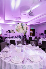 Beautiful table setting with crockery and white flower arrangement in a vase on a high stem for a party, wedding reception or other festive event. Glassware and cutlery for catered event dinner
