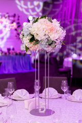 Beautiful table setting with crockery and white flower arrangement in a vase on a high stem for a party, wedding reception or other festive event. Glassware and cutlery for catered event dinner