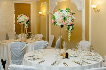 Beautiful table setting with crockery and white flower arrangements in a vase on a high stem for a party, wedding reception or other festive event. Glassware and cutlery for catered event dinner