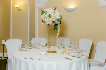 Beautiful table setting with crockery and white flower arrangements in a vase on a high stem for a party, wedding reception or other festive event. Glassware and cutlery for catered event dinner