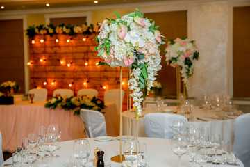 Beautiful table setting with crockery and white flower arrangements in a vase on a high stem for a party, wedding reception or other festive event. Glassware and cutlery for catered event dinner
