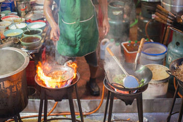 Chef fries in a wok on the vast flame of the stove with rising steam at a street cafe in an alley adjacent to Yaowarat Road, the main artery of Chinatown of Bangkok, Thailand famous with street food