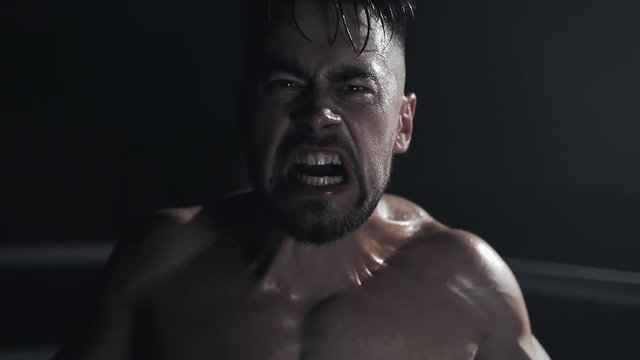 Angry man with boxing gloves screams emotionally looking ar the camera standing on the boxing ring. Slow motion.