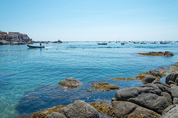 Fototapeta na wymiar A clear and blue morning over the seashore of Calella. A boat arrives to the village among boats and the rocks at foreground.