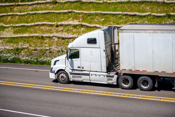White big rig semi truck with dry van semi trailer driving on the road with hill on the background