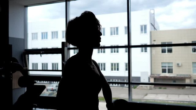 Silhouette of a sportive woman with a relief body at the window making set of reps exercise lifting barbell for shoulders muscles.