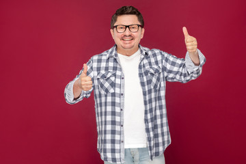 Naklejka premium Portrait of happy excited middle aged business man in casual checkered shirt and eyeglasses standing, thumbs up, toothy smiling looking at camera. indoor studio shot, isolated on dark red background.
