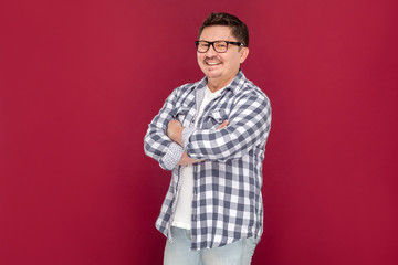 Portrait of successful middle aged business man in casual checkered shirt and eyeglasses standing, crossed arms and looking at camera with toothy smile. studio shot, isolated on dark red background.