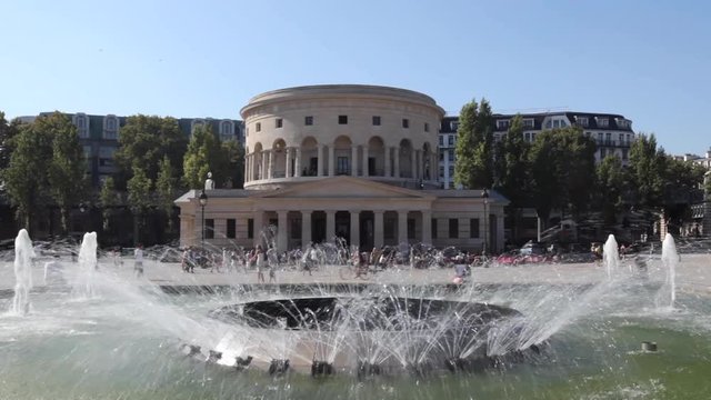 Paris, France - April 5, 2019: Rotonde de La Villette with fountain in the foreground and lots of people walking down the street. Blue sky background.