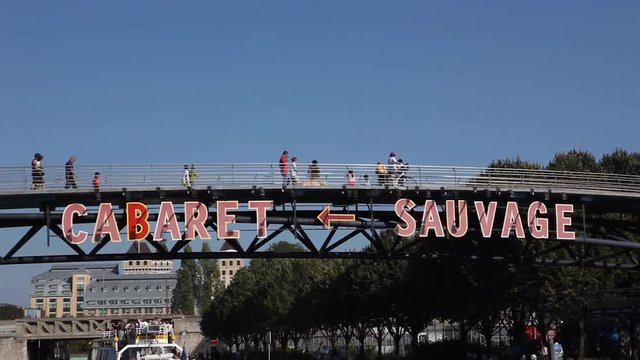 Paris, France - April 5, 2019: Parisian boat passing under a bridge at the Parc de la Villette with the inscription Cabaret Sauvage. People crossing the bridge. Sunny summer with blue sky. Tight shot.