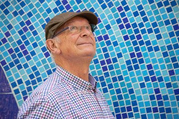 Retrato de un hombre mayor con gafas, boina y camisa, mirando al cielo con un mosaico de fondo.