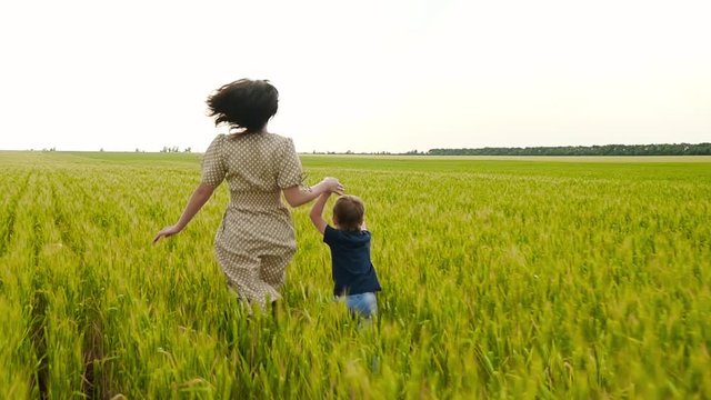 A young mother and her child run across a field of wheat, holding hands. The concept of a happy family, motherhood and agriculture.