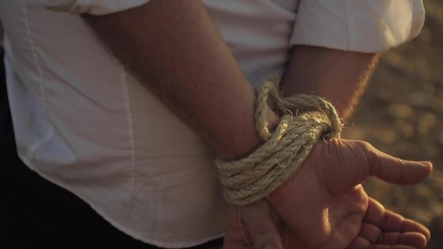 Hands of man tied in the back by a thick rope in the Moroccan desert in the middle of a heat wave during the sunny summer. Man standing still, kidnapped by criminals. Sun rays on the hands. Close up.
