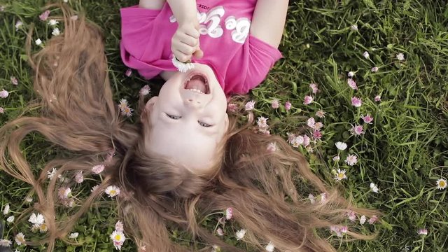 Close-up smiling little cute baby girl lying on green grass holding flowers looking at camera upside down view. Face of happy pretty young kid enjoying nature at summer day