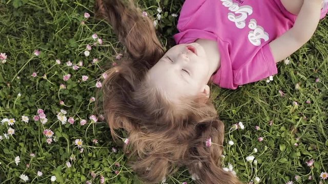 Close-up smiling little cute baby girl lying on green grass holding flowers looking at camera upside down view. Face of happy pretty young kid enjoying nature at summer day