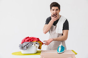 Young confused displeased houseman househusband with iron near clothes isolated over white wall background talking by mobile phone.