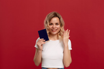 Portrait of a girl with curly blond hair dressed in a white t-shirt standing on a red background. Model smiles at the camera, holds passport of blue color and lifts hands with a gesture of surprise