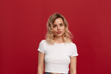 Portrait of a beautiful girl with curly blond hair dressed in a white t-shirt standing on a red background.