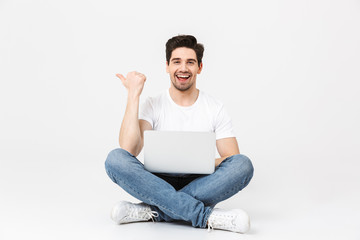 Excited young man posing isolated over white wall using laptop computer pointing sitting on floor.