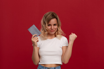 Portrait of a girl with curly blond hair in a white t-shirt standing on a red background. Model smiles at the camera, holds passport and expresses gesture of success.