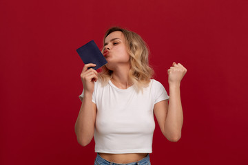 Portrait of a girl with curly blond hair in a white t-shirt standing on a red background. Model smiles at the camera, holds passport and expresses gesture of success.